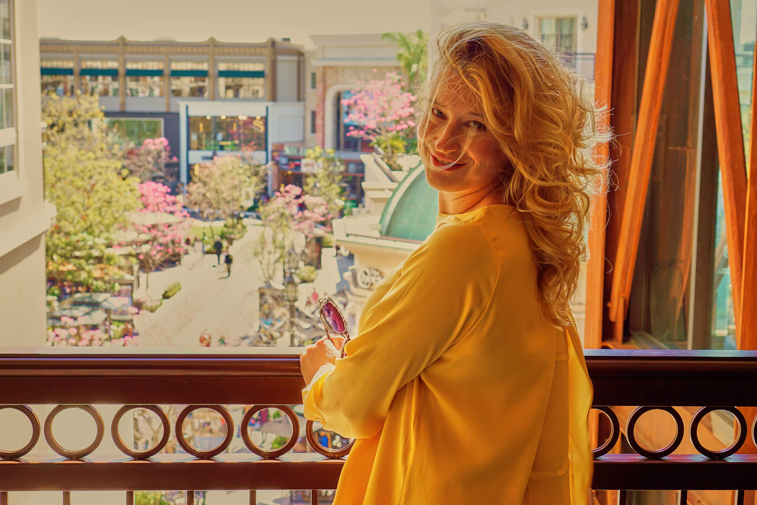 A woman in a mustard-yellow blouse stands on a balcony, turning back with a smile while holding sunglasses, overlooking a shopping courtyard with pink flowering trees.